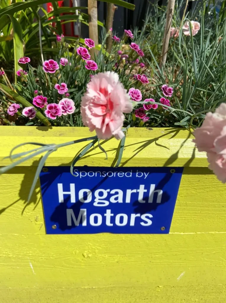 A yellow painted planter of pink flowers with a sponsor plaque
